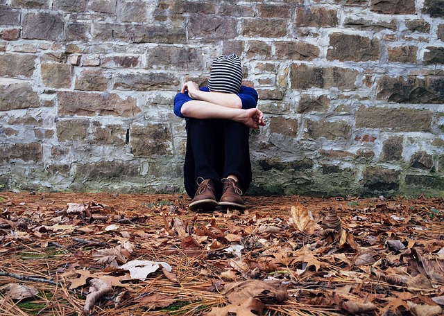 A sad teen, sitting against a wall outside, with their arms folded across their knees and their head down.