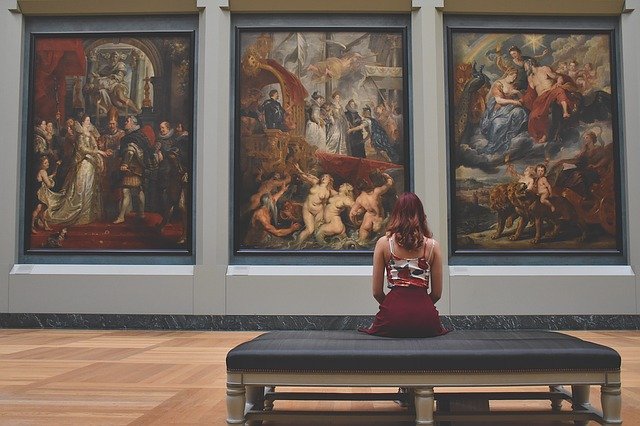 A woman sitting on a bench in a museum, looking at three large framed paintings.